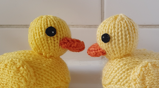 two yellow knitted rubber ducks facing each other with plain white bathroom tiles in the background