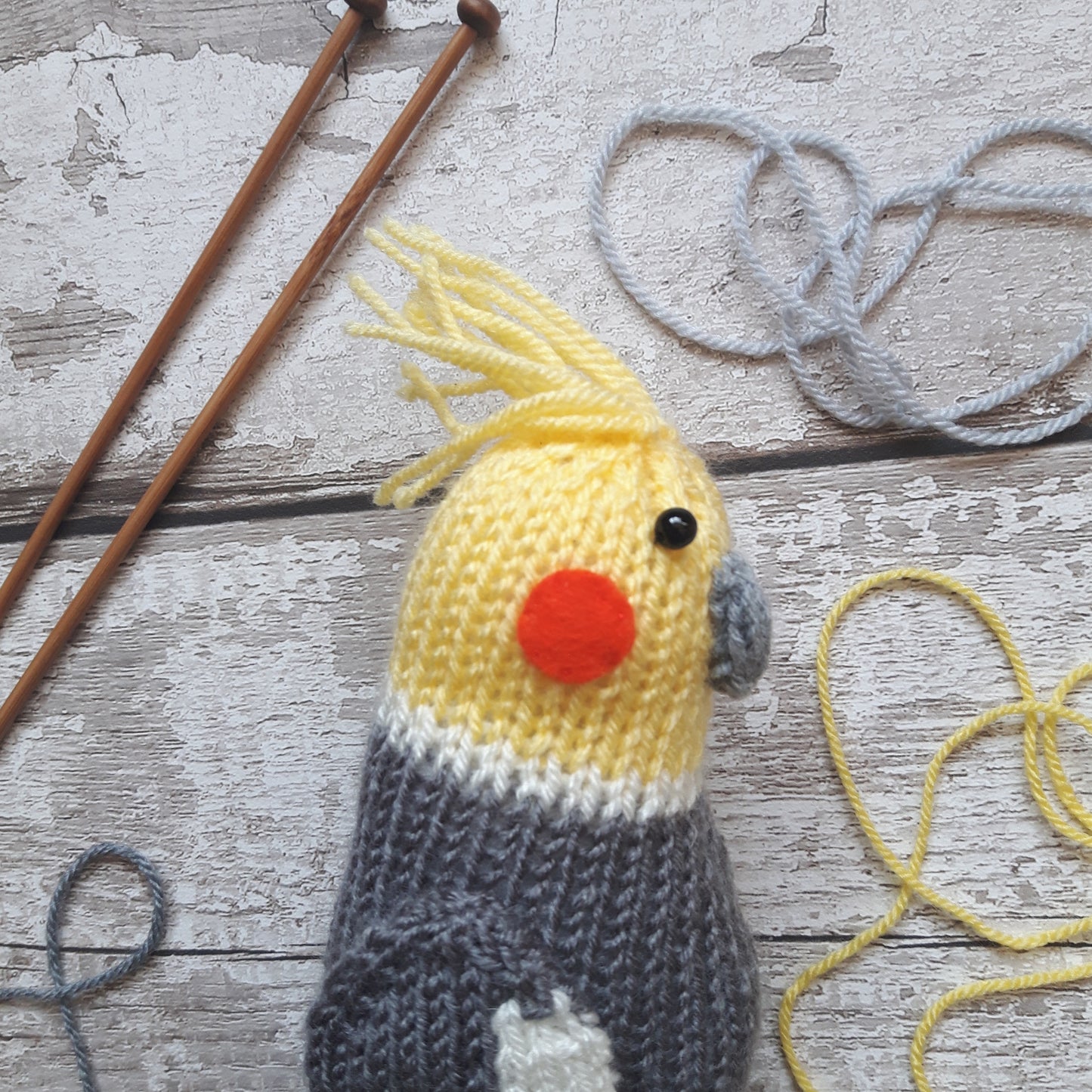 a grey and yellow knitted cockatiel with a pair of wooden knitted needles and grey and yellow yarn in the background