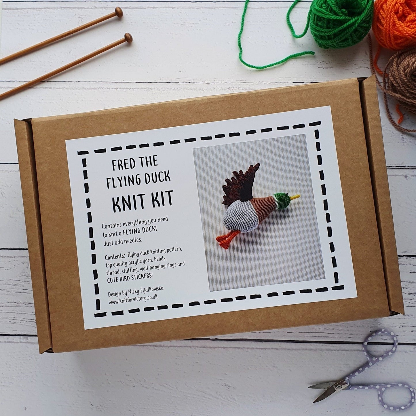 A knitting kit for a flying duck, in a brown card box. In the background there are knitting needles, scissors and some colourful yarn