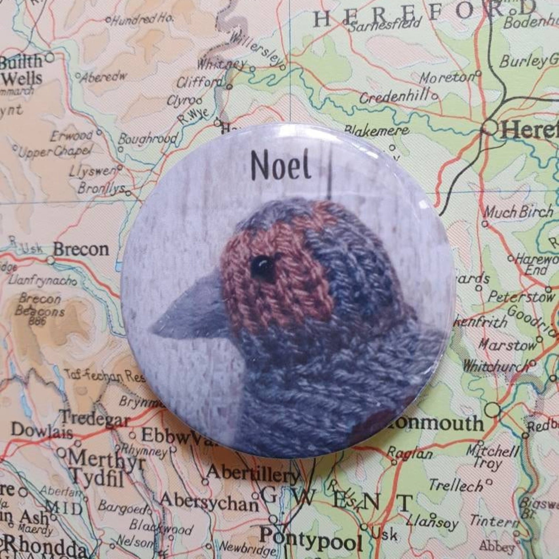 a noel the partirdge button badge with the word Noel on it and a photo of a knitted partridge. The badge is sitting on a vintage map.