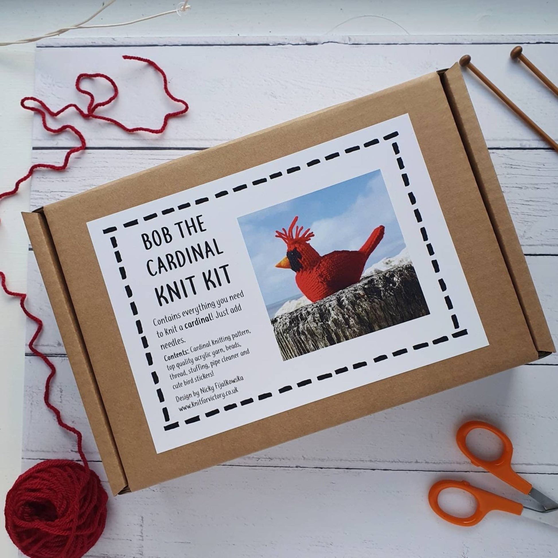 a northern cardinal knit kit in a box. In the background is scissors and red yarn