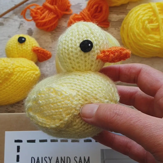 a yellow knitted rubber being held. In the background you can see yellow yarn, a knit kit and knitting needles