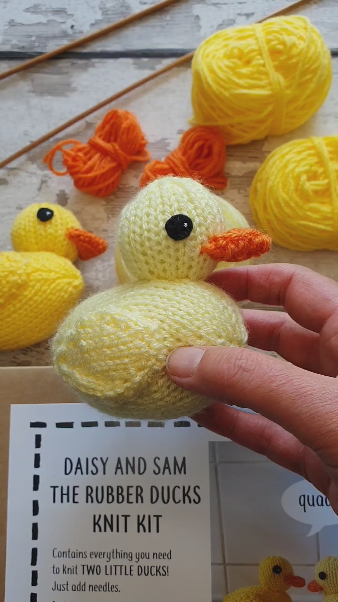 a yellow knitted rubber being held. In the background you can see yellow yarn, a knit kit and knitting needles