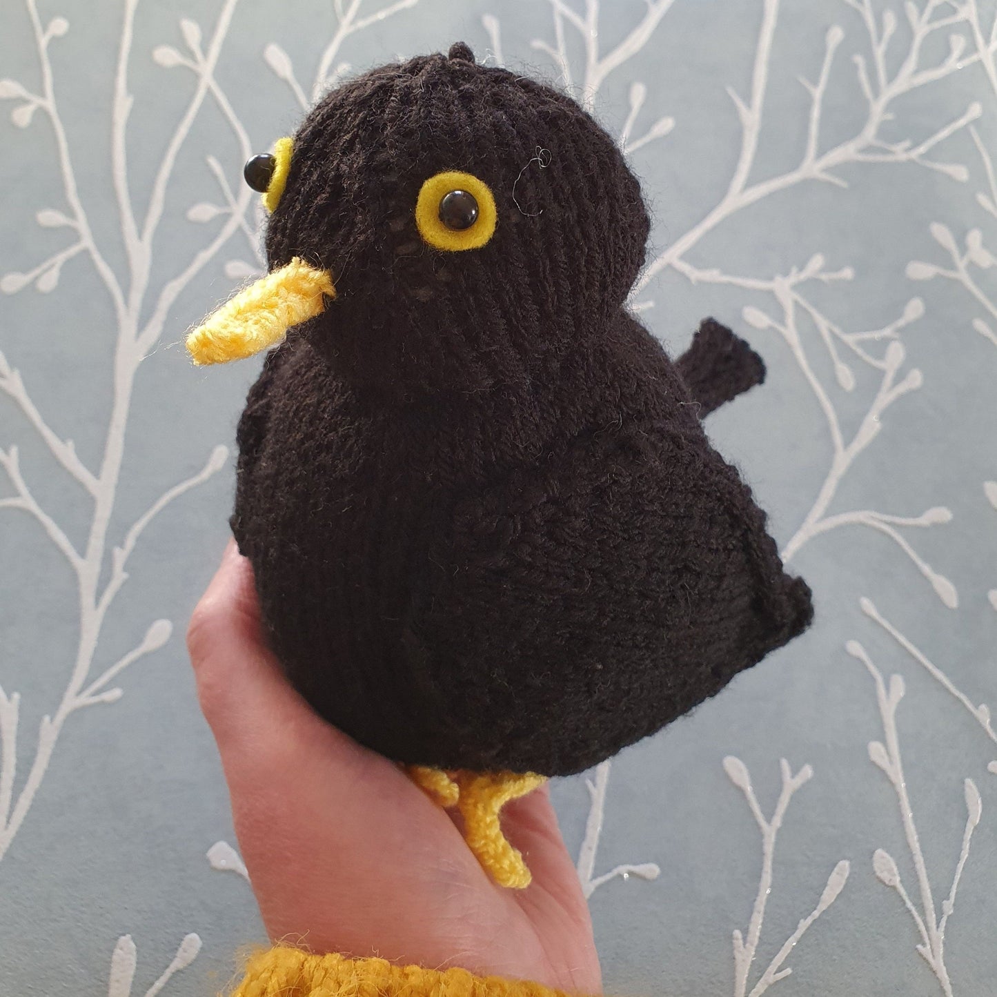 a plump knitted blackbird being held in a female hand. There is grey wallpaper in the background.